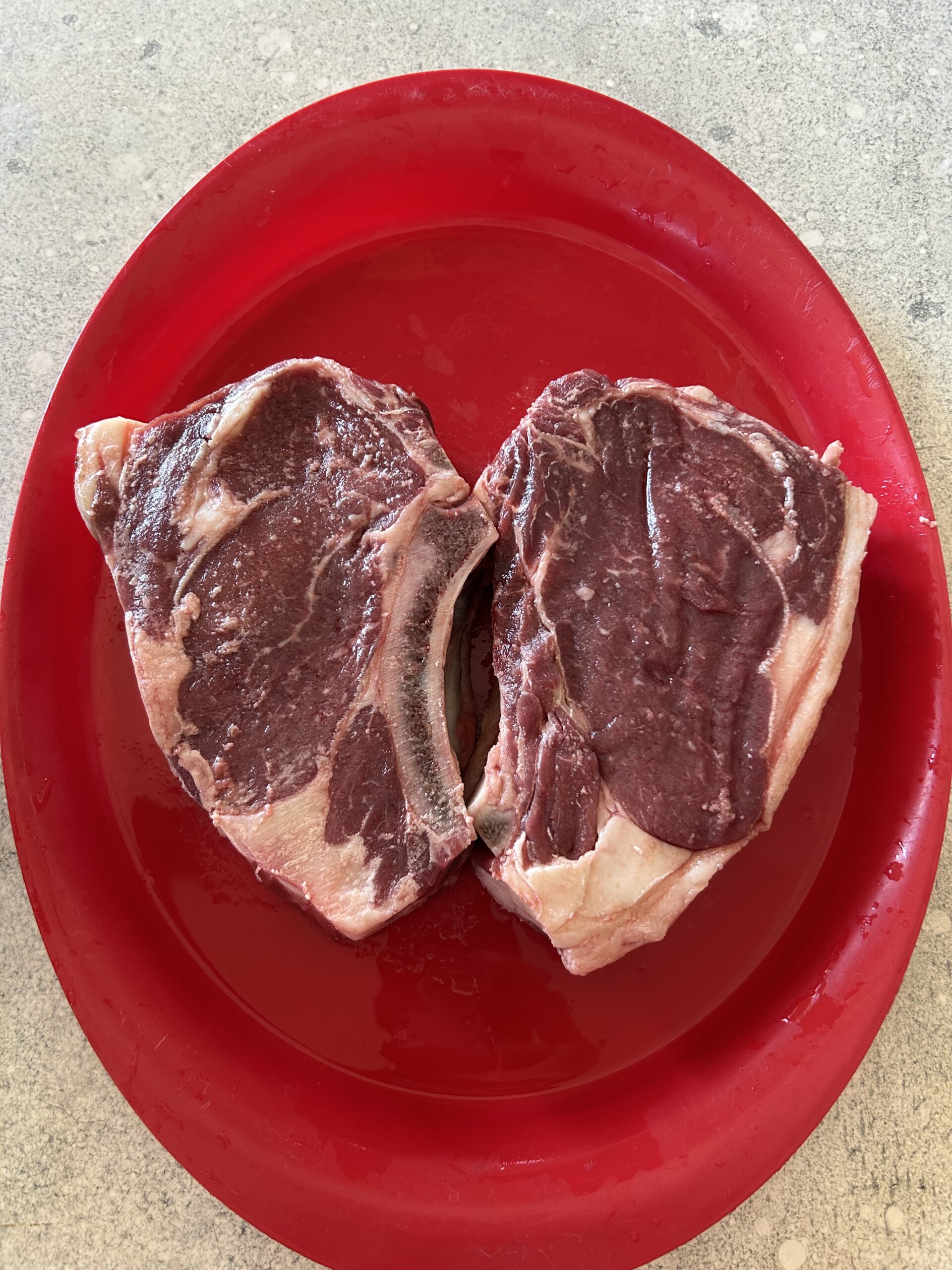 Two raw ribeye steaks displayed on a red plate, showcasing marbling and fat content, ready for grilling or cooking.