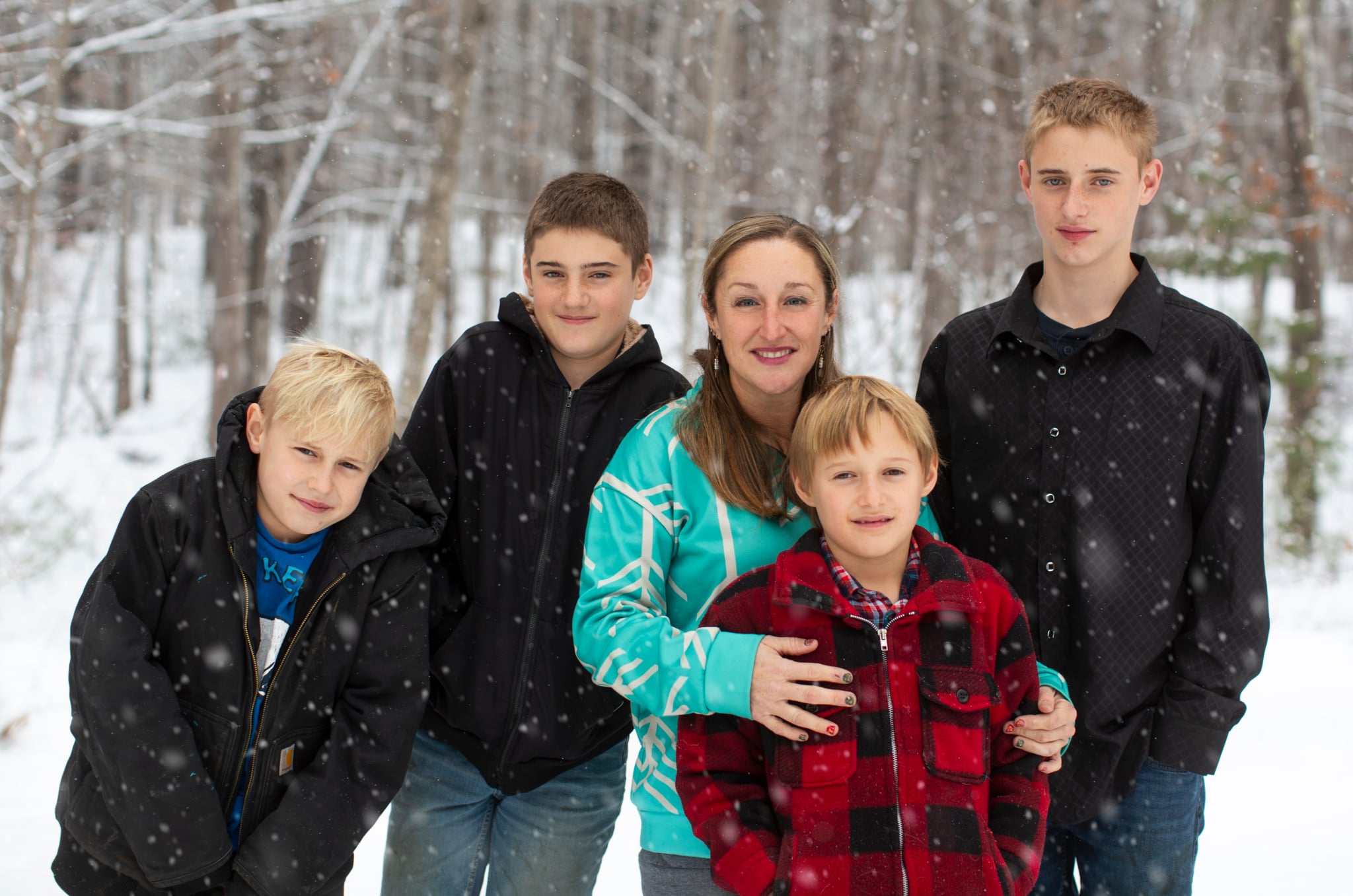 A mother and her four sons pose together in a snowy forest, wearing winter jackets. The scene captures a joyful family moment amidst falling snowflakes, showcasing the beauty of winter and family bonding.